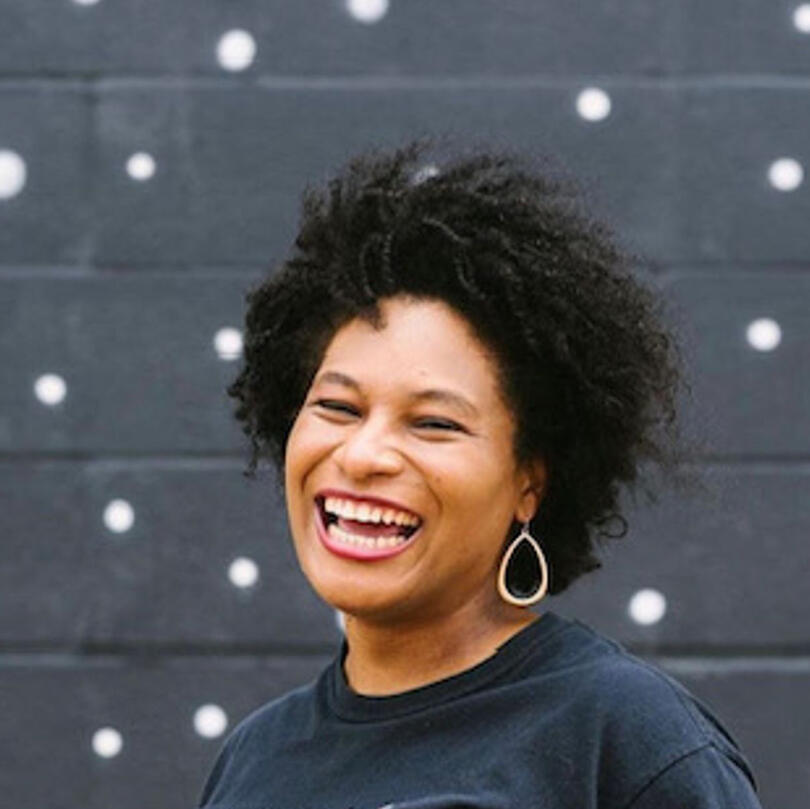 Image of Ashley Borjon smiling. She is wearing gold teardrop-shaped earrings and a black shirt. She stands in front of a black brick wall with white circles.
