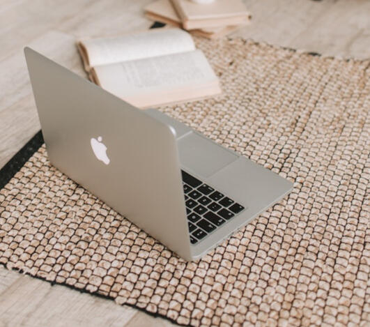 Image of a silver MacBook and three books, with one opened. The books and laptop are on top of a light brown handwoven rug.